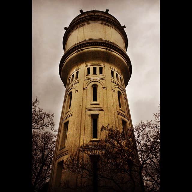 Torre del agua de ladrillo, fotografiada desde abajo contra un cielo claro y rodeada de ramas desnudas, en tonos sepia.