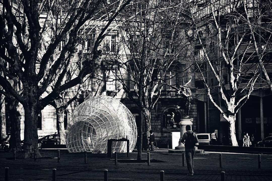 Plaza arbolada con una escultura esférica de metal y un peatón, en blanco y negro.