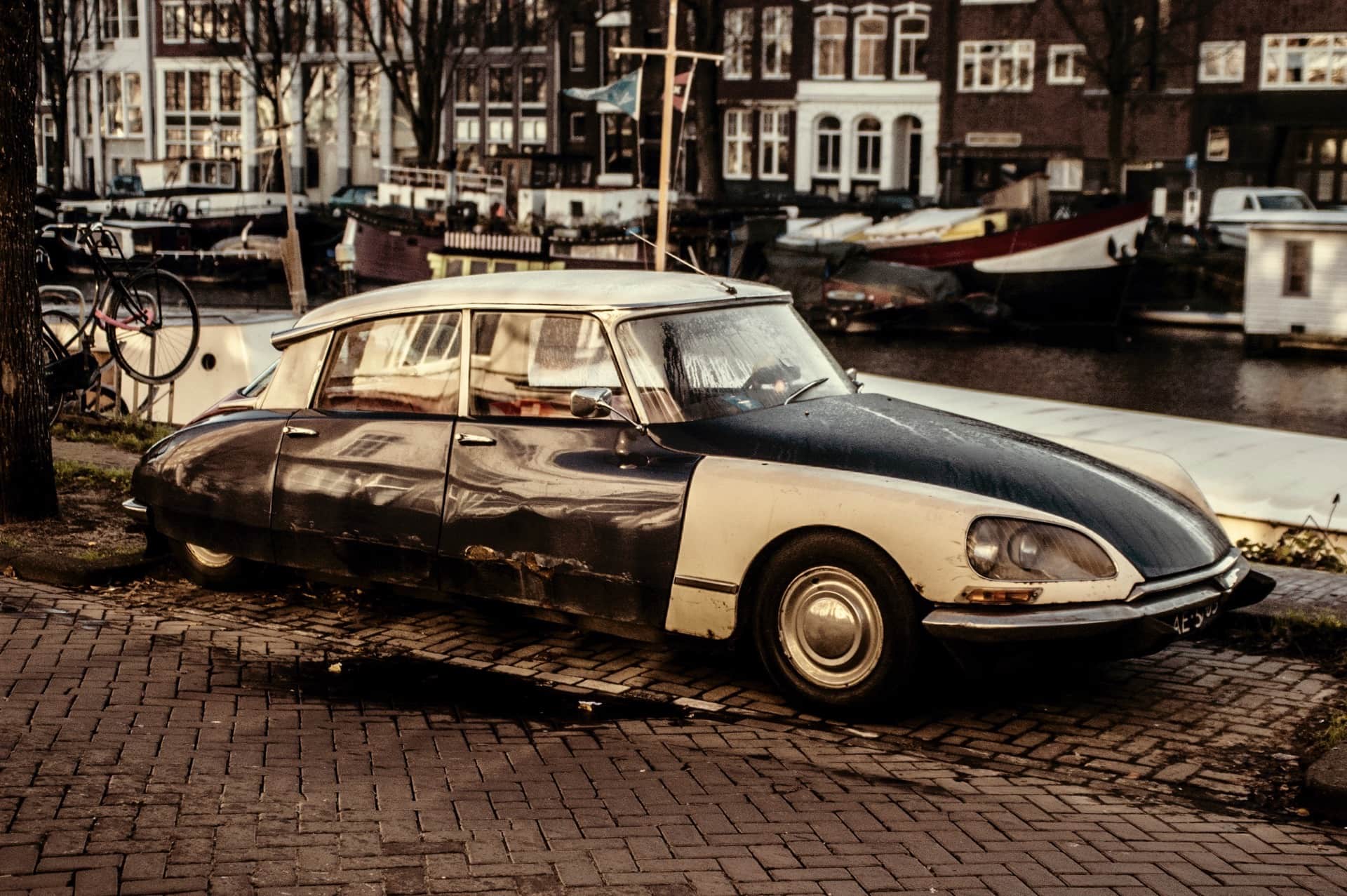 Citroën DS bicolor con la pintura desgastada, aparcado en un muelle de ladrillo frente a un canal con casas flotantes.