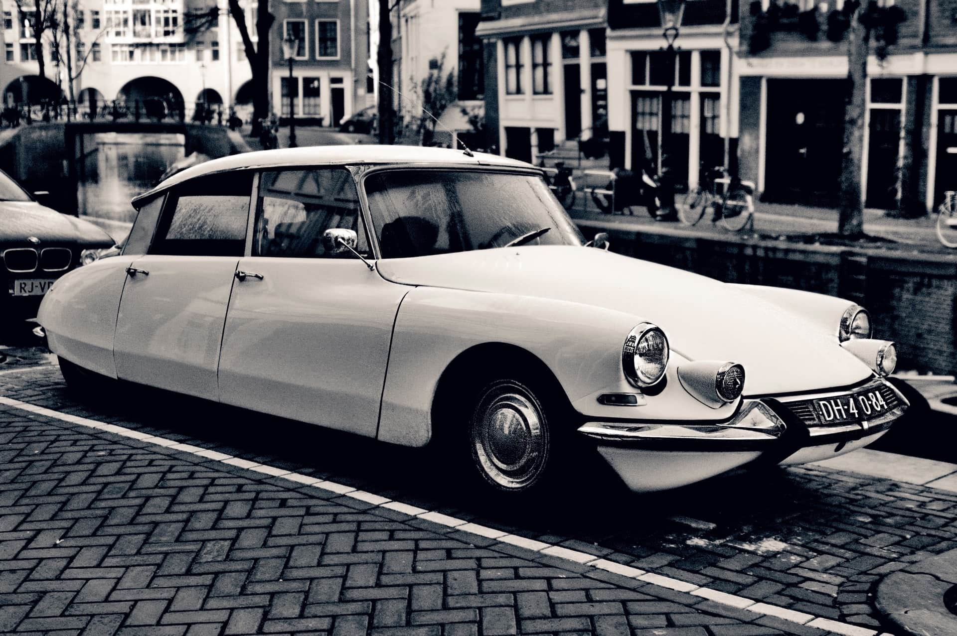 Citroën DS claro aparcado sobre adoquines junto a un canal, con casas estrechas y un puente al fondo, en blanco y negro.