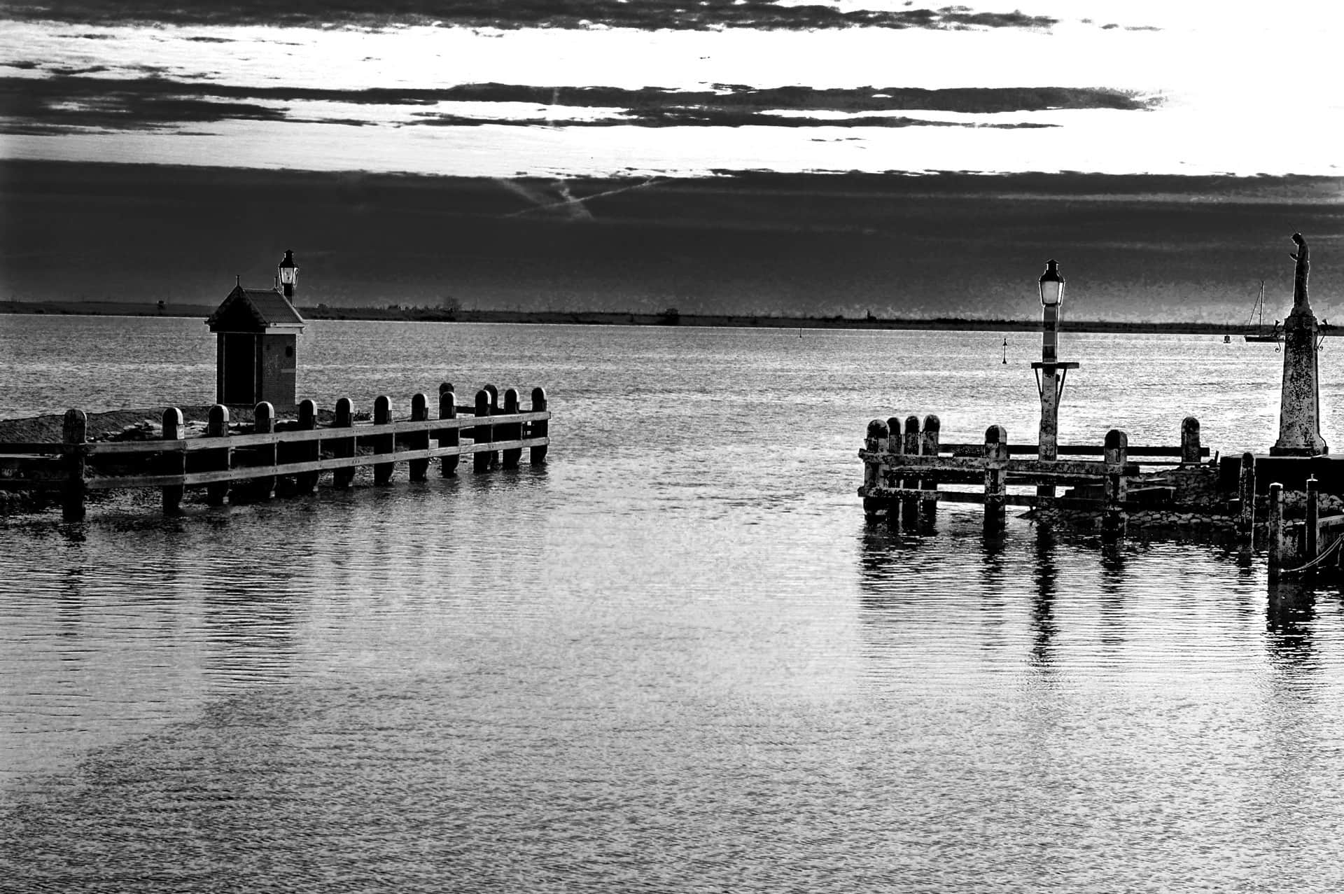 Espigones de madera sobre agua en calma, con una caseta de ladrillo con farol y una estatua sobre un pilar de piedra, en blanco y negro.