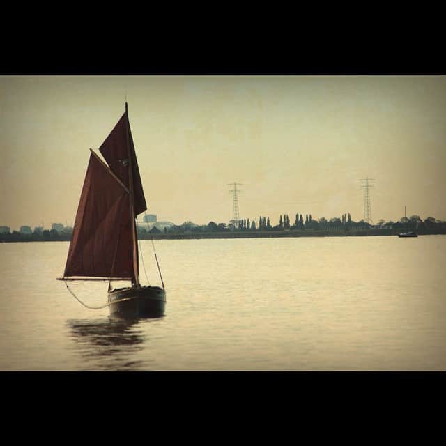 Velero de vela oscura navegando en un agua muy calma, con la orilla arbolada al fondo, en tonos sepia.
