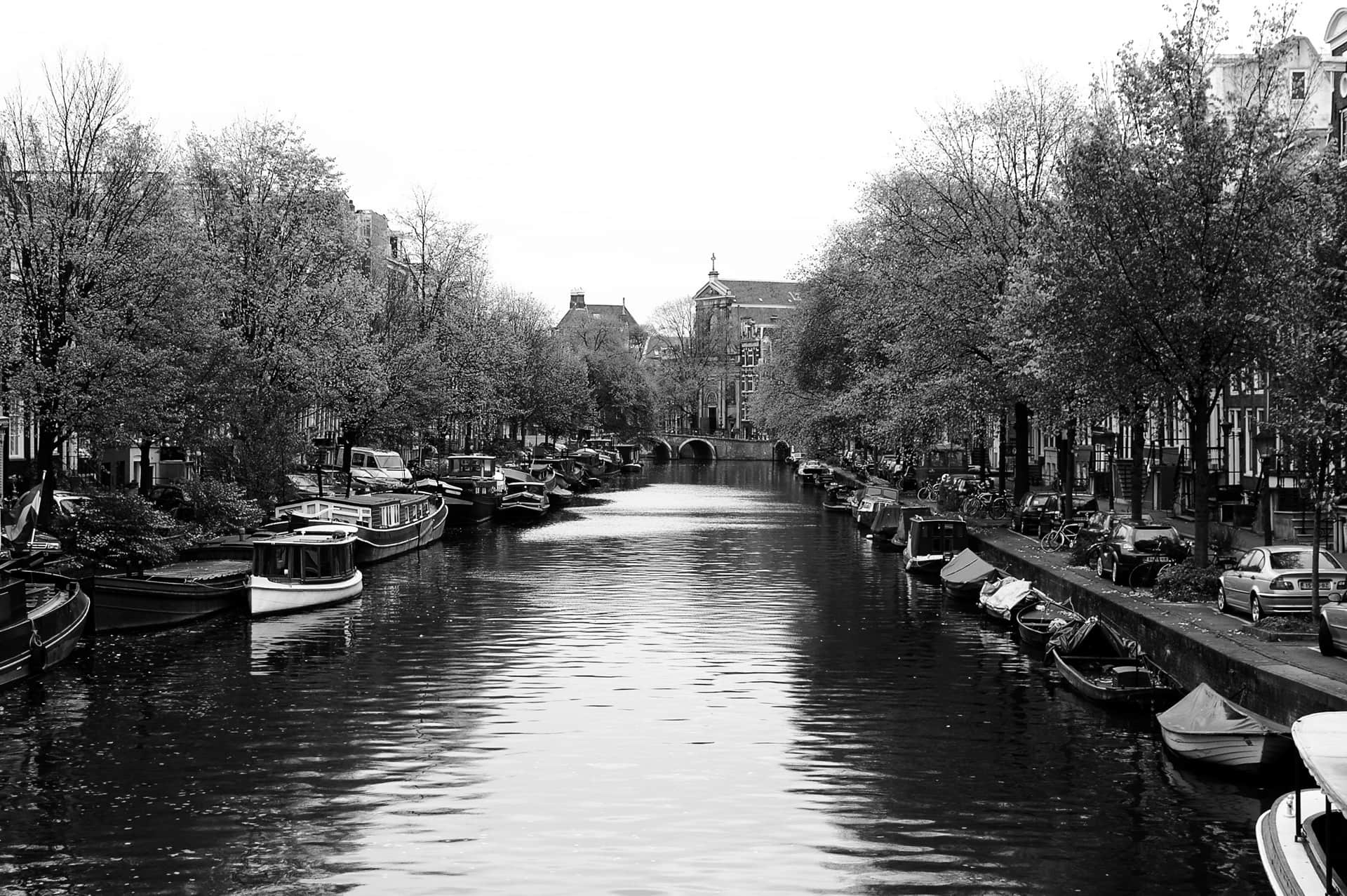 Canal urbano flanqueado por árboles y barcazas amarradas, con un puente de arcos de ladrillo y una iglesia al fondo, en blanco y negro.