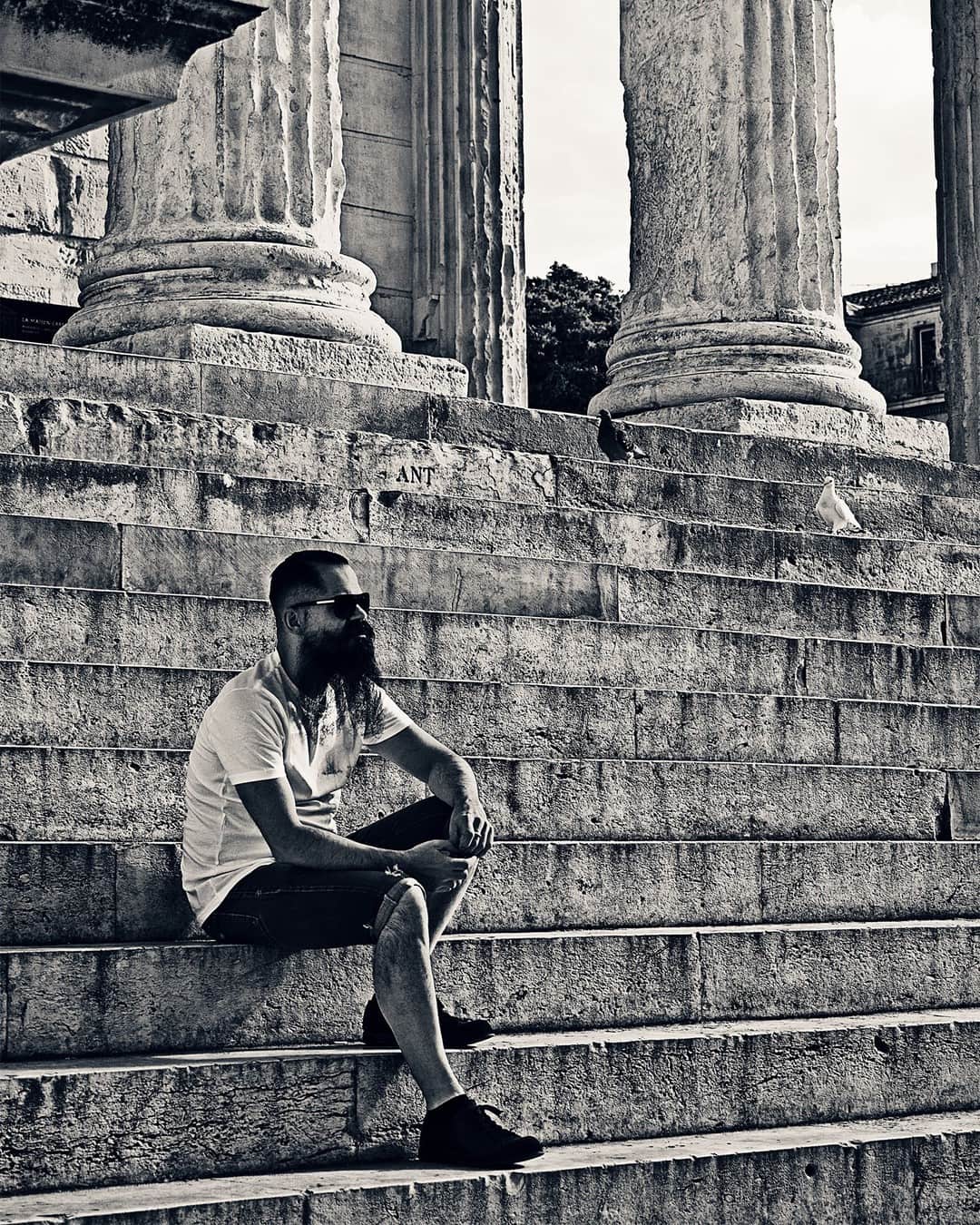 Hombre joven sentado en una escalinata de piedra junto a unas columnas, en blanco y negro.
