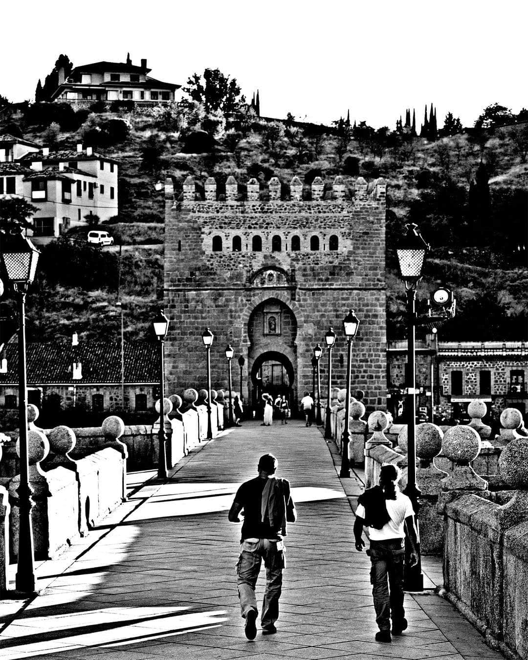 Puente de Toledo con dos hombres caminando hacia la puerta fortificada, en blanco y negro.