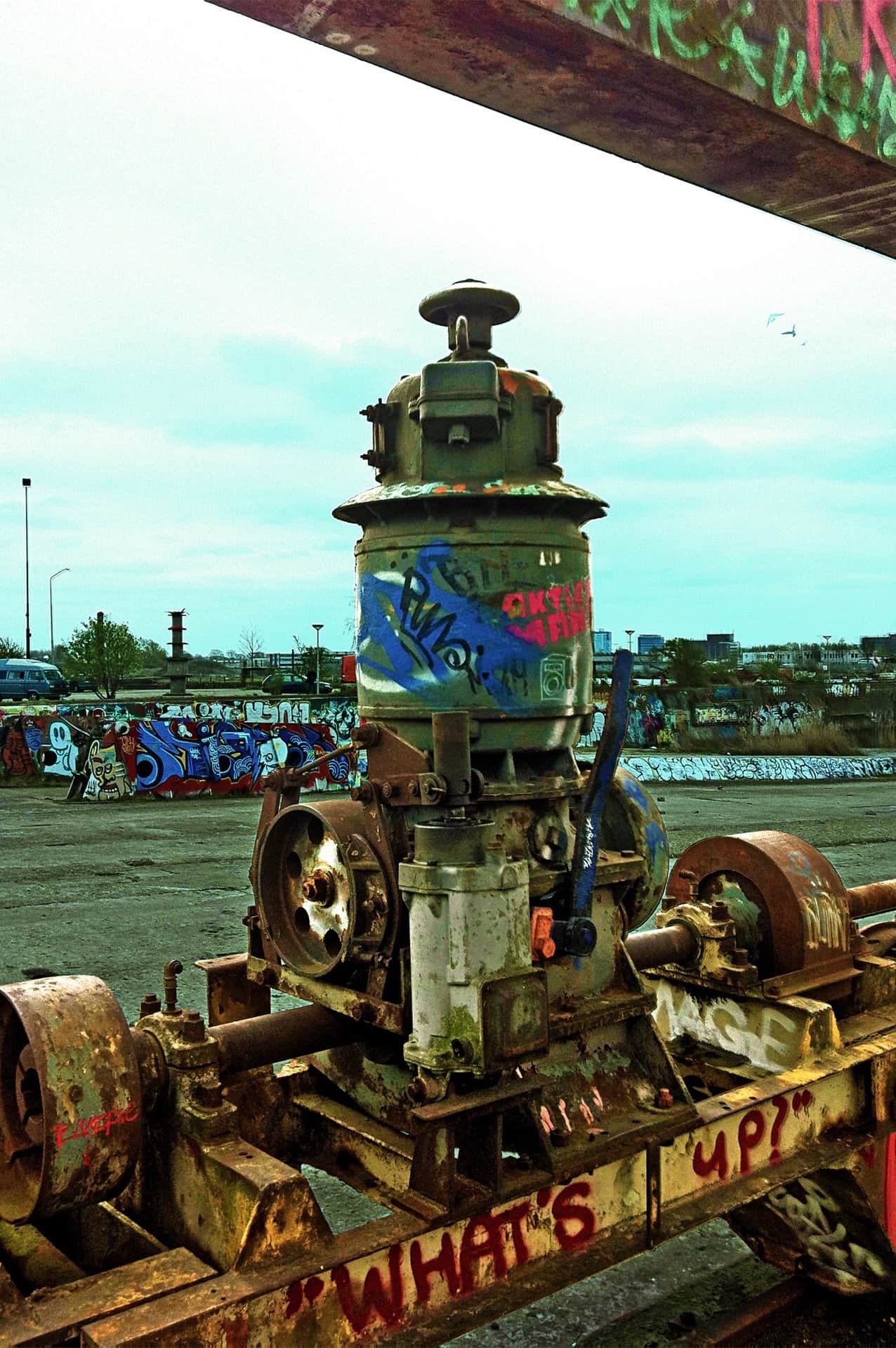 Maquinaria industrial oxidada y cubierta de grafitis en una explanada de hormigón, con «WHAT'S UP?» pintado en rojo en el bastidor.