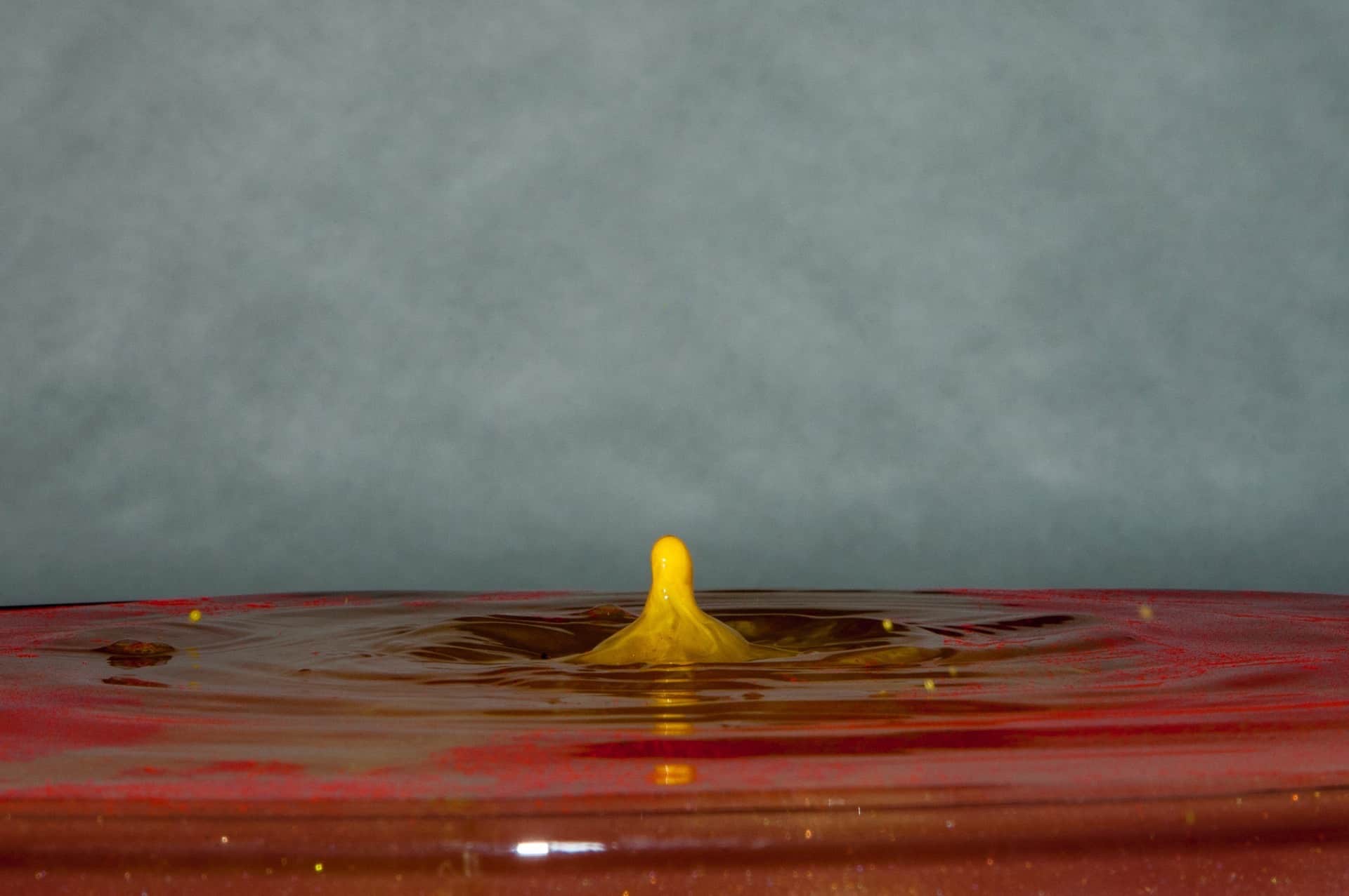 Columna de líquido amarillo alzándose tras el impacto de una gota en una lámina rojiza, sobre fondo gris liso.