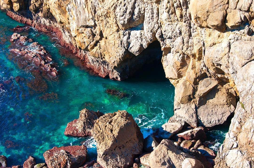 Cala de agua turquesa entre rocas claras, con la boca oscura de una cueva al fondo.