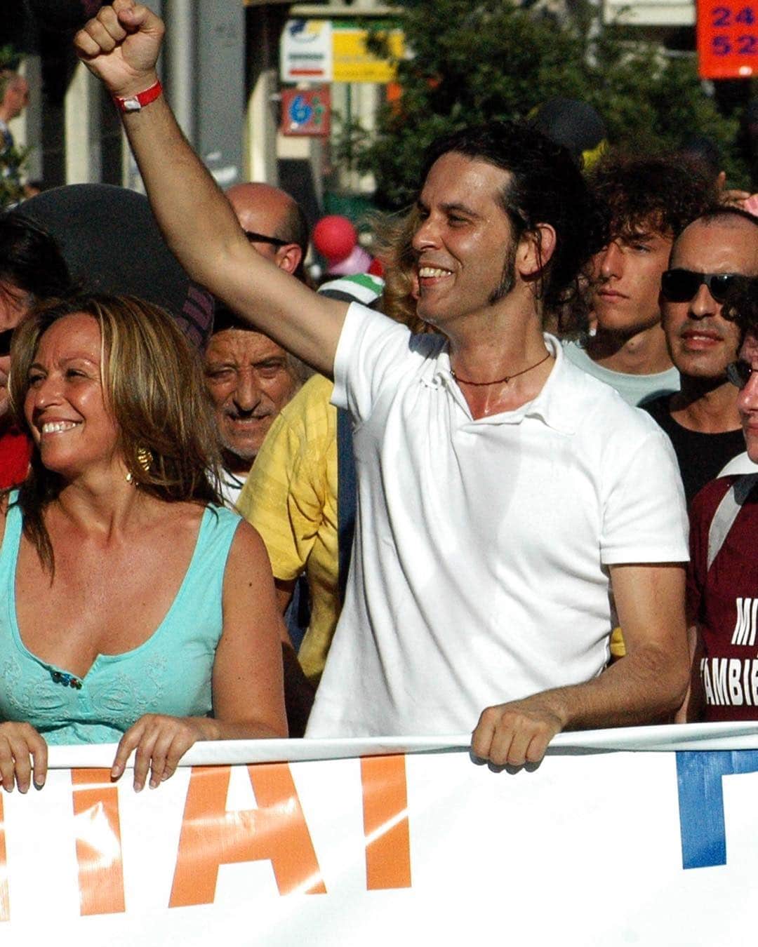 Hombre con el puño en alto y camiseta blanca, sonriendo entre la multitud de una manifestación, detrás de una pancarta.